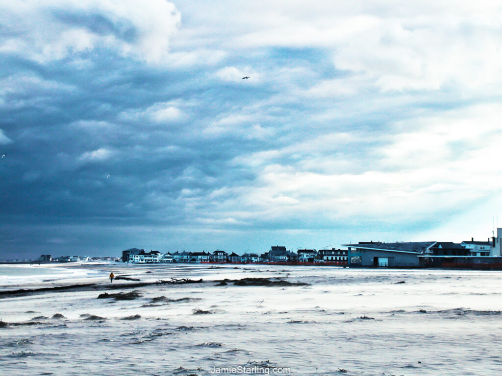 Quiet winter beach in Hampton, NH, with a lone figure walking along the shoreline, under expansive cloudy skies—evoking a sense of solitude and warmth.