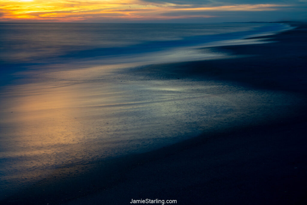 A serene beach at sunset with golden light reflecting on wet sand and deep blue ocean tones, capturing the harmony between light and dark, stillness and motion.