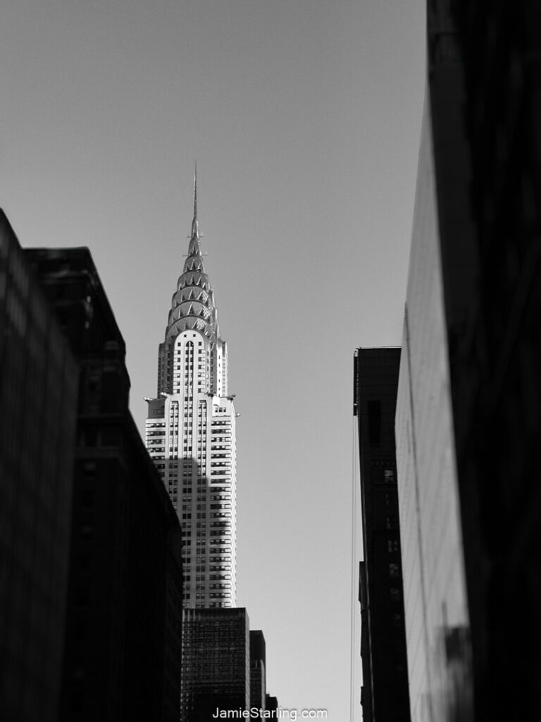 Black and white photograph of the Chrysler Building, standing tall and striking against a minimal backdrop with shadowed buildings, symbolizing elegance and individuality.