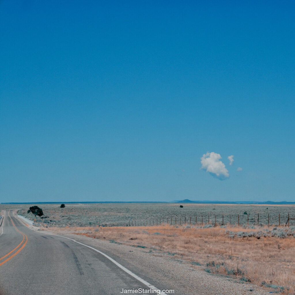 A winding road stretching into the horizon under a clear blue sky, with a single cloud floating above a barren yet expansive landscape—an image of simplicity and stillness inviting reflection.